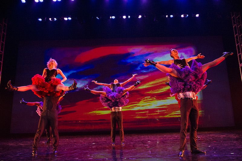 Women in ruffly red skirts are lifted above their male partners heads with their legs out-stretched